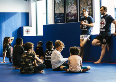 Young kids sitting on a blue mat with coaches standing talking to them